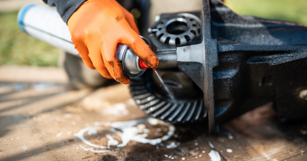 A close-up of a person applying a cleaner from an aerosol can to a car differential on top of a piece of cardboard.