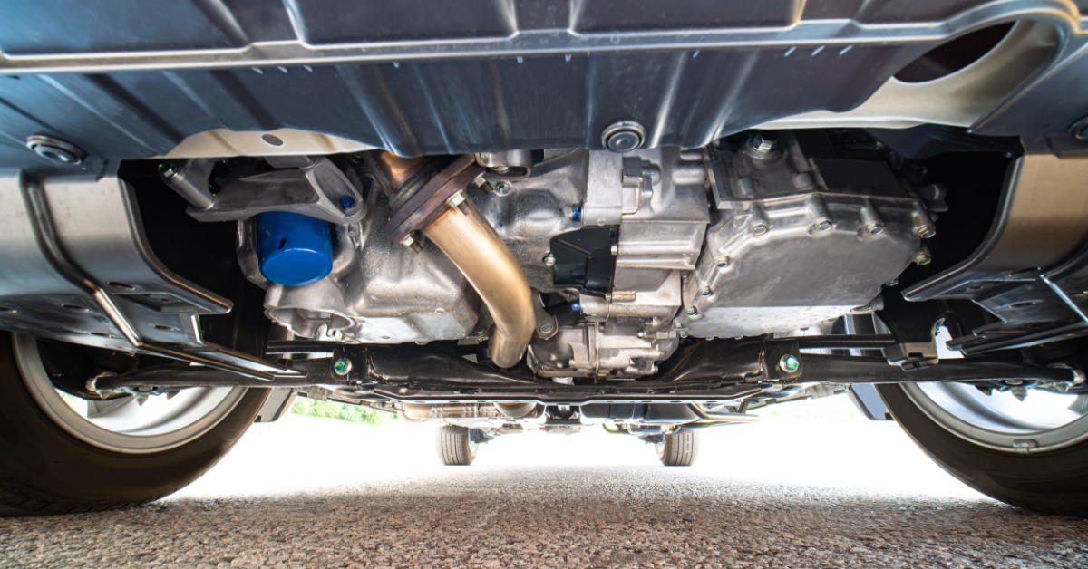 The underside of a vehicle with a blue oil filter, curved exhaust pipe, metal pans, and front tires above the pavement.