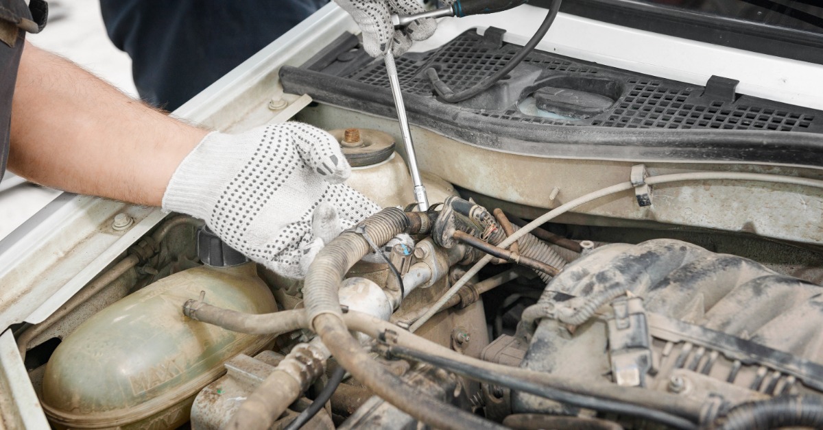 A person wearing gloves uses a wrench on hoses and metal parts inside the engine compartment of a vehicle.
