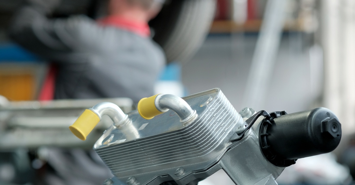 A silver transmission oil cooler with yellow tube caps and a black cylinder rests on a workbench in an auto shop.