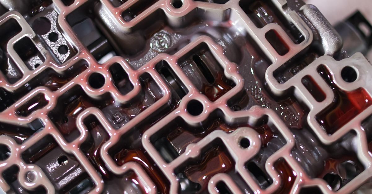A metal transmission valve body with rectangular channels and round holes covered in red hydraulic fluid.