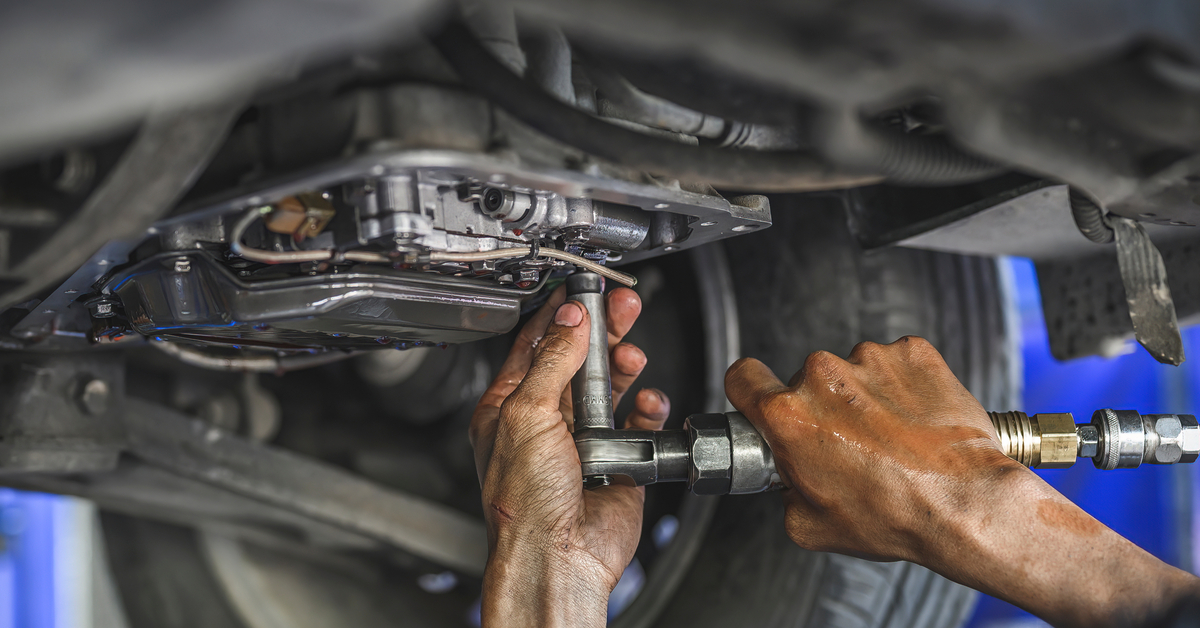 An elevated car. A mechanic tightens a bolt on the drivetrain. His hands are slick and dirty from grease and oil.