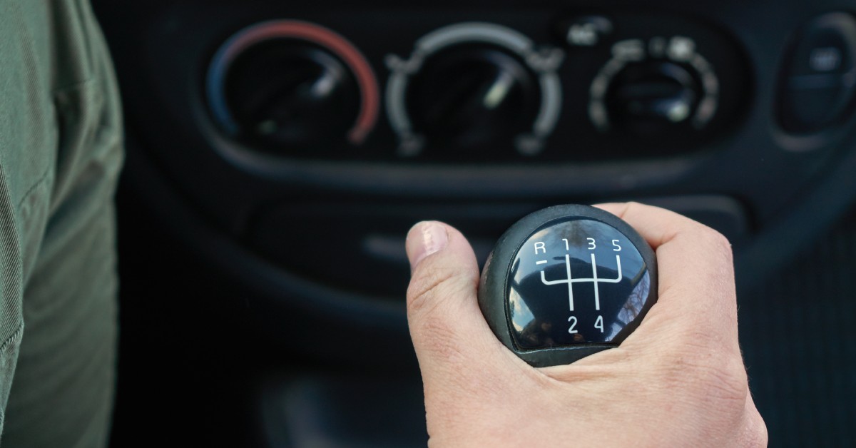 A hand grips the gearshift of a manual car. The climate control knobs are out of focus in the background.