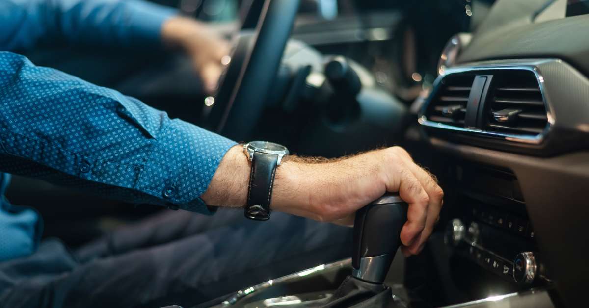 A man's hand shifts the automatic transmission in his car. He wears a watch and a blue, long-sleeved shirt.