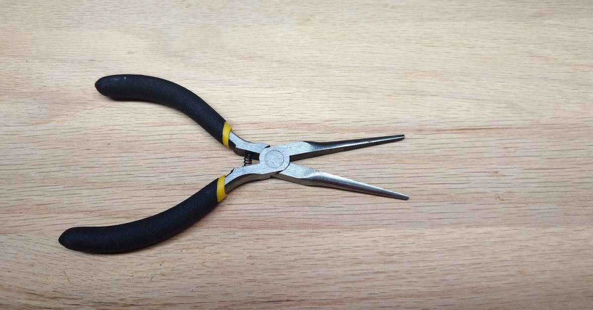 A pair of dirty needle-nose pliers with black and yellow handles sits on a wooden surface. The pliers have a spring.