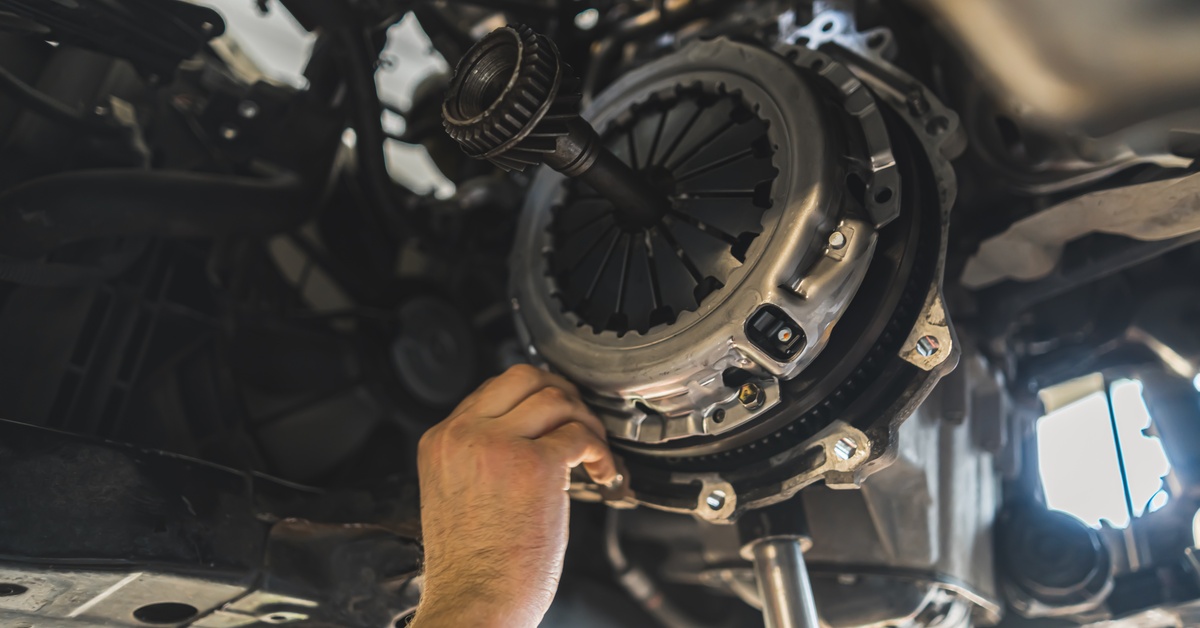 A close-up of a hand installing a transmission clutch on the underside of a vehicle. Various gears and other parts are visible.