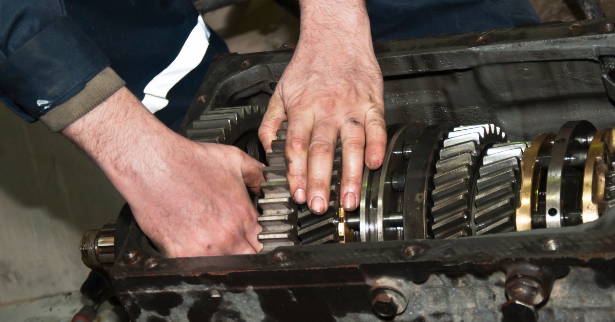 Close-up of a person's hands working on a car transmission. The person wears a dark blue jumpsuit that has white stripes.