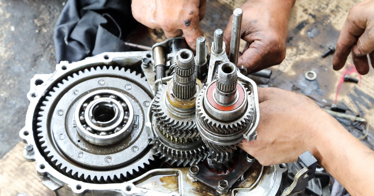 Two parts of hands work on repairing a car's gear box. The gear box sits on cardboard on the floor of a garage.
