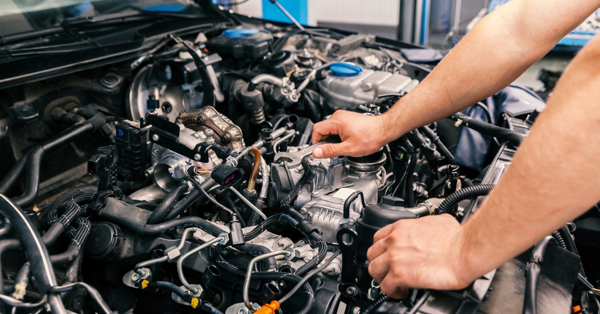 A person works on the engine of a vehicle while holding a small wrench in one hand. The person has a red-sleeved shirt.