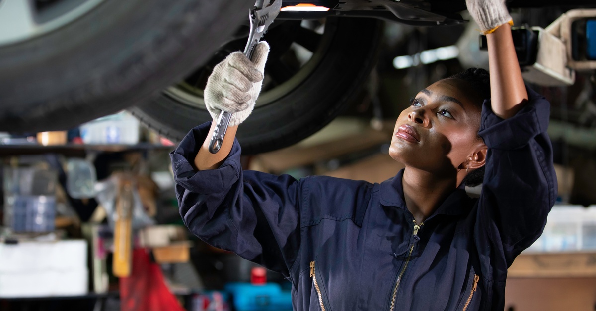 A woman wearing a blue jumpsuit works on a car that is on lifts in her garage. She holds a wrench in her hand.