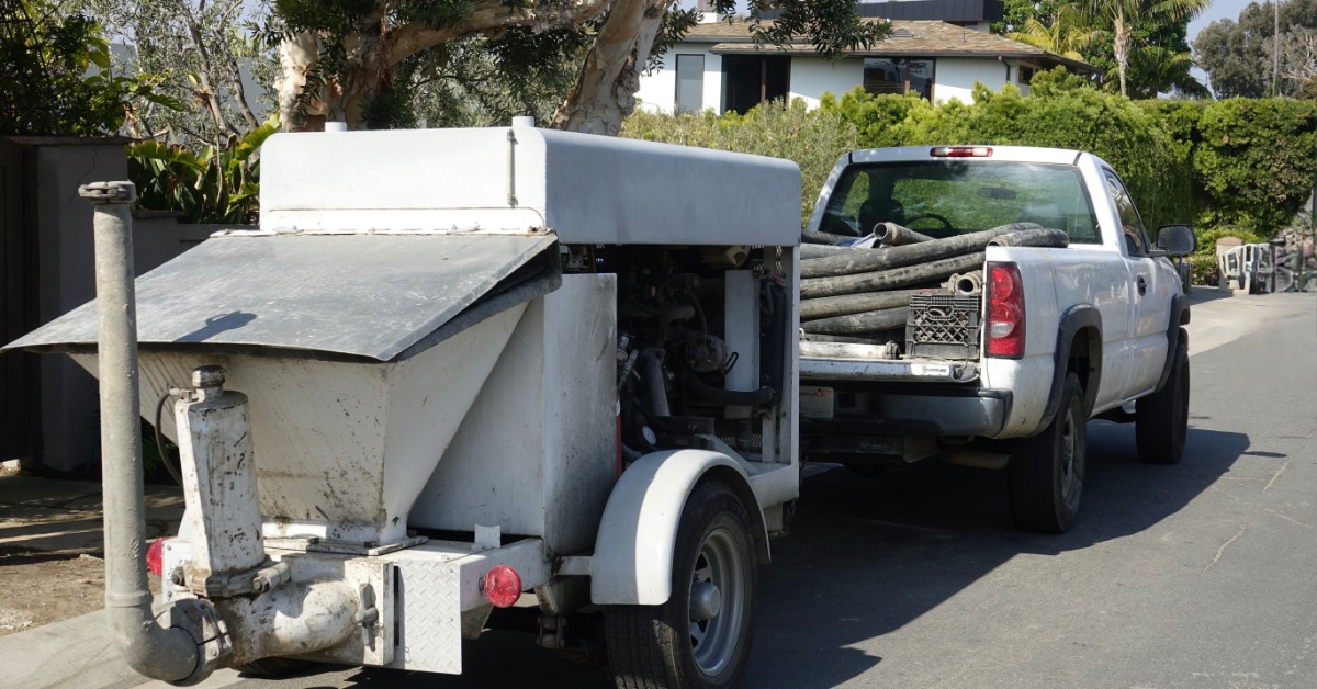A pick-up truck tows a concrete pumper as it drives along a road. Several pieces of pipe are in the back of the truck.