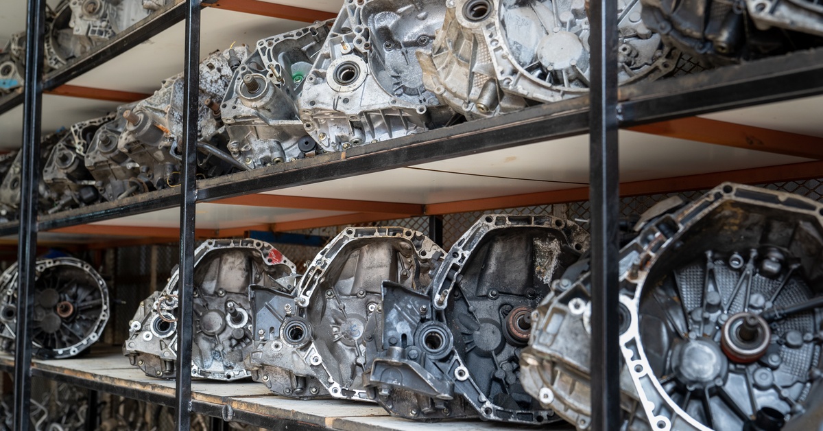 Transmission parts sit on tall shelves in a storeroom. The shelves have black metal frames and wood panels.