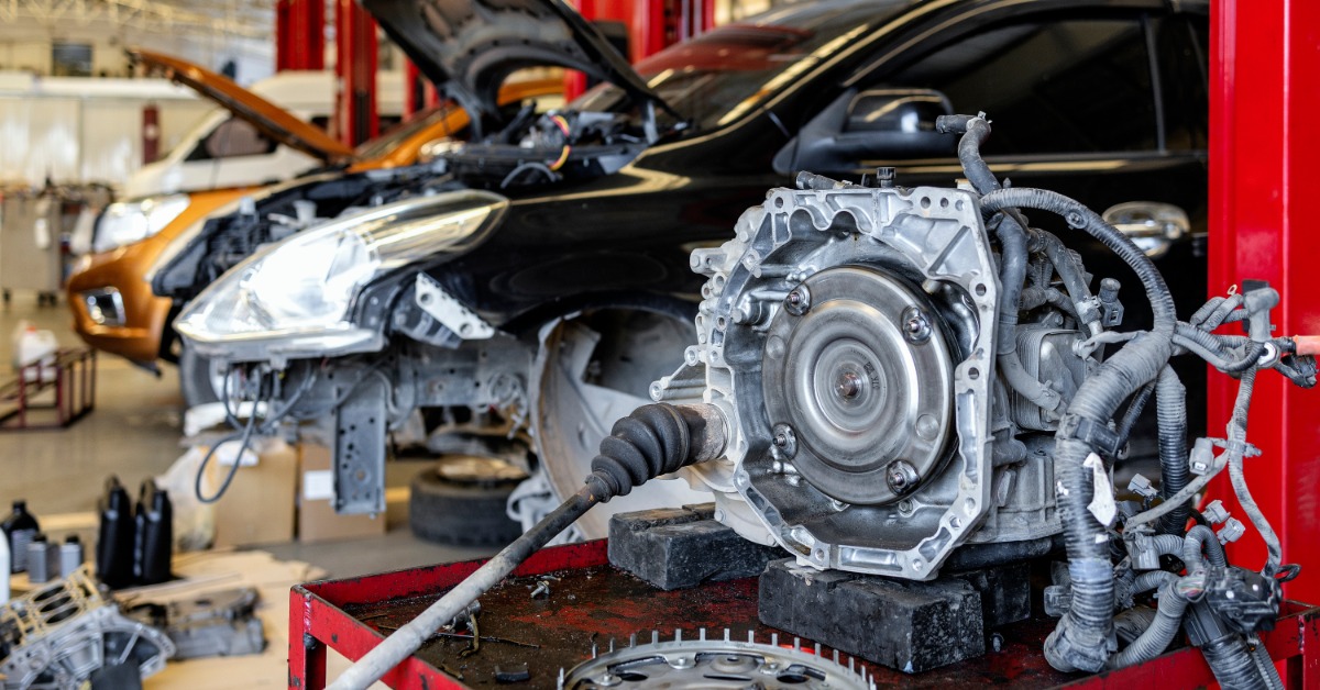 A disassembled automatic transmission rests on a workbench with parts in a garage. A raised car sits behind it.