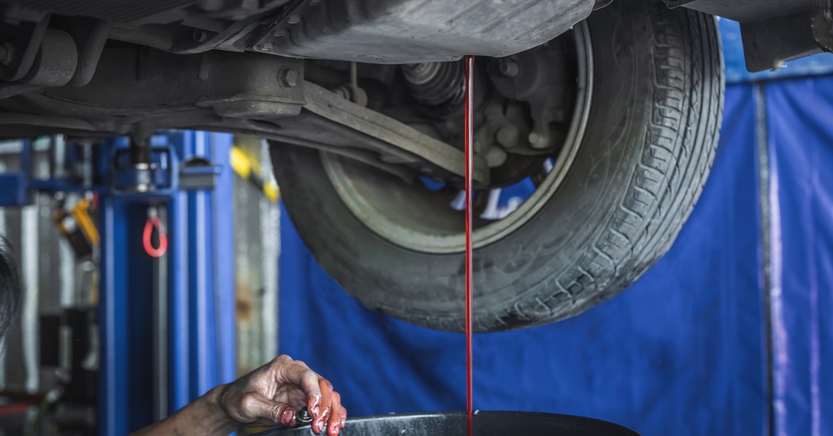 Red fluid drains from a car's transmission pan into a container. A hand holds a plug below the vehicle.