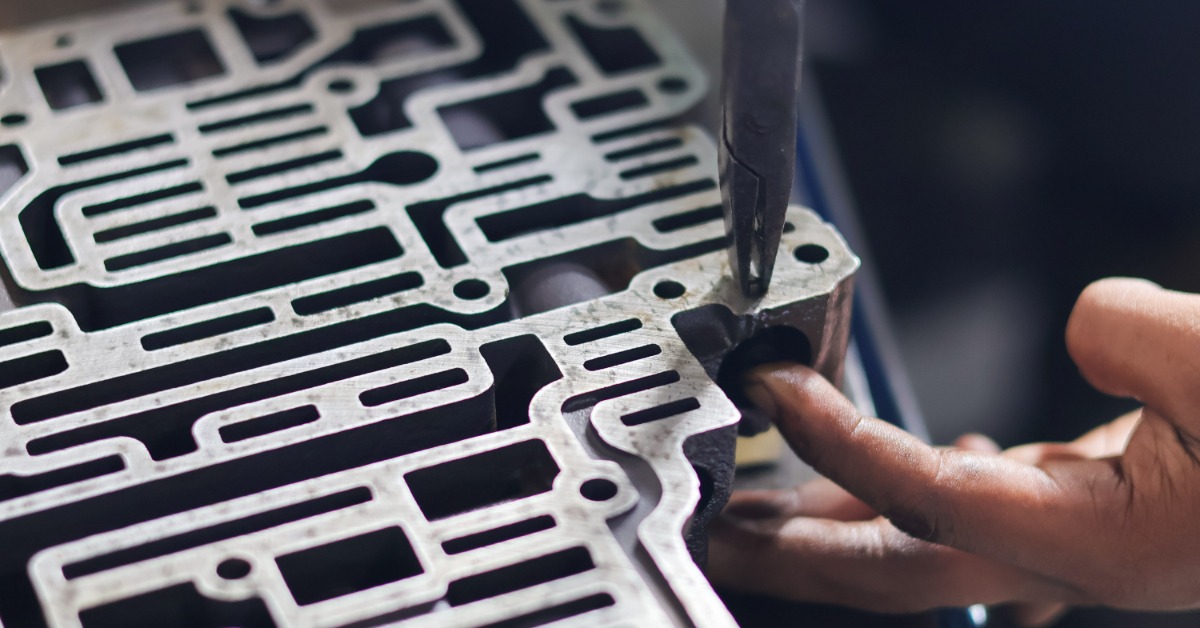 A hand guides a pointed tool along narrow channels and round openings on a metal transmission valve body.