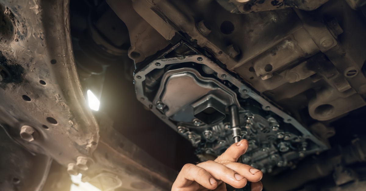 Hands using a socket tool beneath a vehicle on an exposed transmission part with bolts and metal casing.