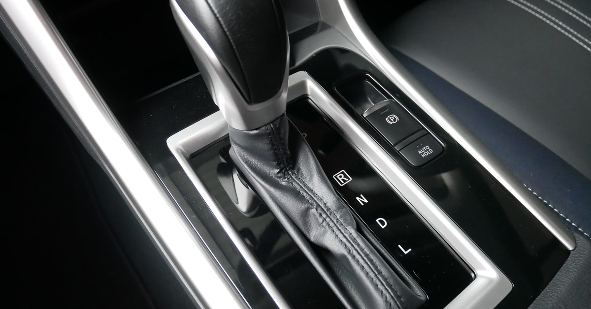 Close-up of a gear shift stick in a car, with the various gears next to it in white letters. It sits near a gray car seat.