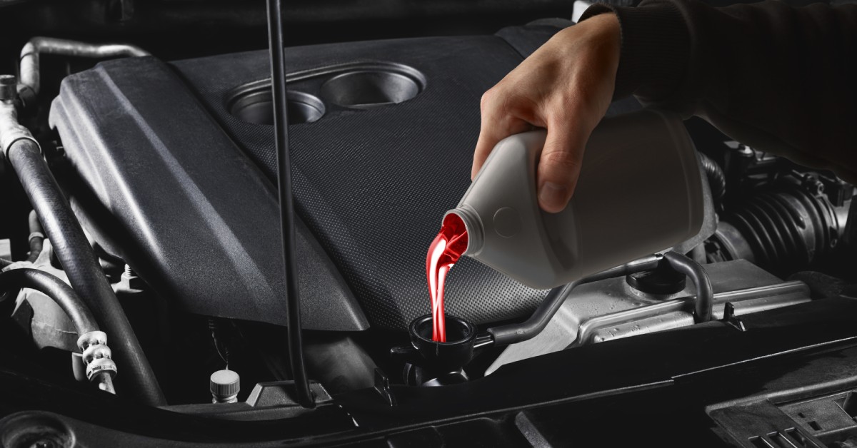A person's hand holds a white container, which pours bright red transmission fluid into a car's engine.
