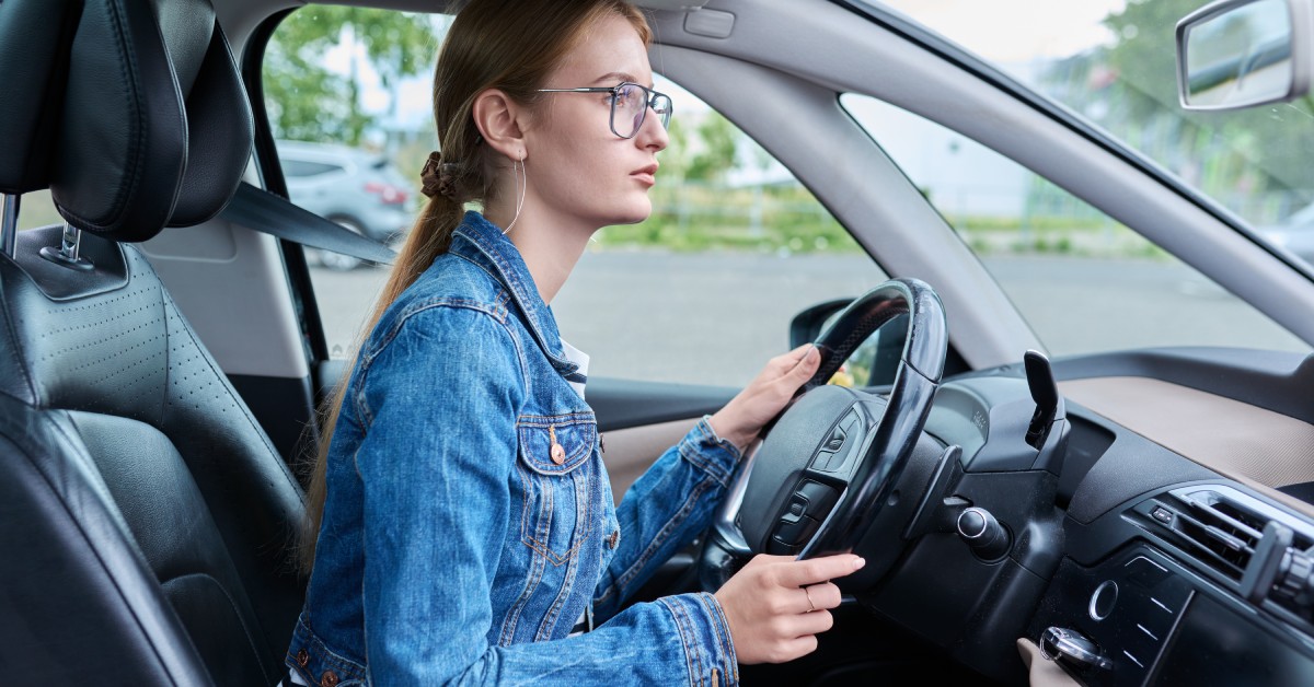 A woman looks carefully as she sits behind the wheel of her car. She wears a denim jacket and glasses.