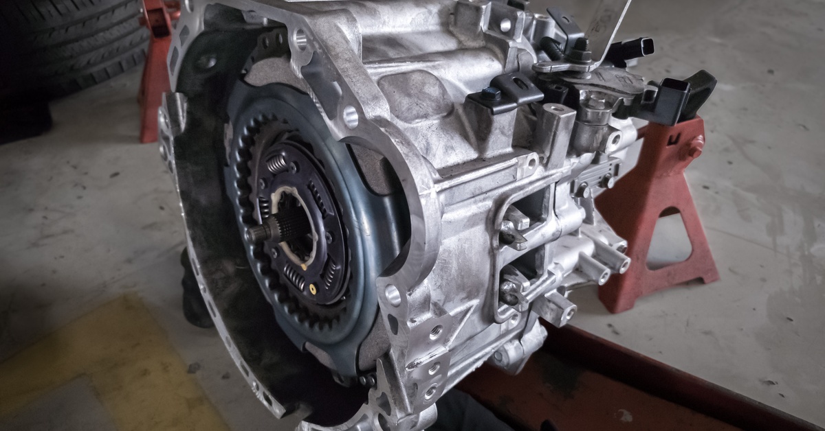 A silver automatic transmission sits on a jack stand on a garage's floor. A car's wheel sits nearby.