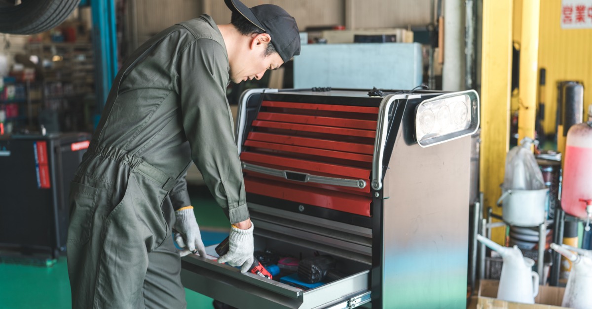 A mechanic wearing a jumpsuit and a baseball cap looks through his tools while standing in his garage.