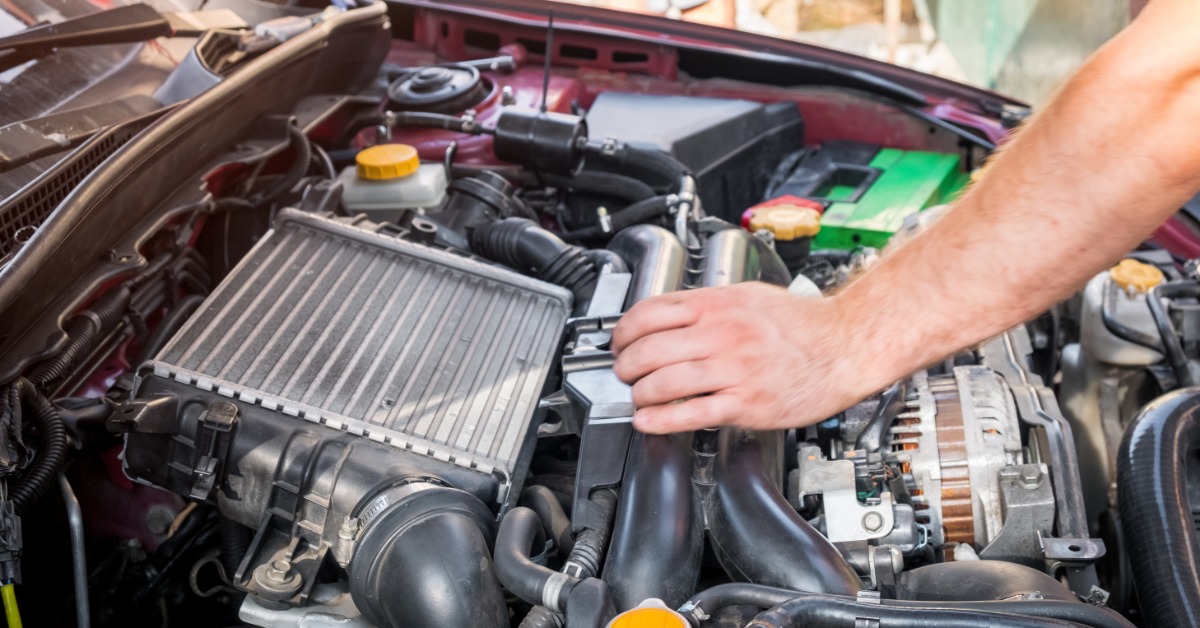 Close-up of a person's hand working underneath the hood of their car. An intercooler is visible in the engine.